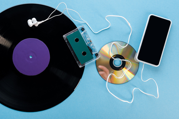 κορυφαία άποψη της vintage βινυλίου ρεκόρ, συμπαγής δίσκος, ακουστικά, ταινία ήχου και smartphone με λευκή οθόνη στο μπλε, έννοια της εξέλιξης  - Φωτογραφία, εικόνα