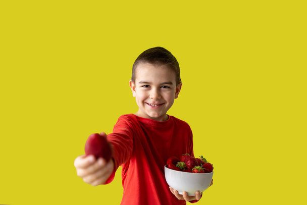 Sonriente niño sostiene una fresa en las manos, cinco años de edad, fondo amarillo, vista frontal
 - Foto, Imagen