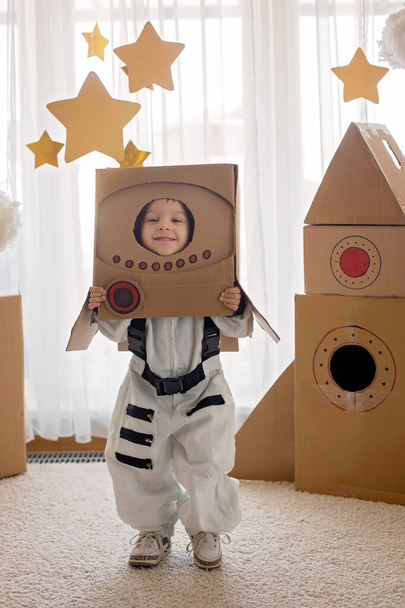 Γλυκό αγοράκι, ντυμένο αστροναύτη, παίζει στο σπίτι με χάρτινη ρόκα και χειροποίητο κράνος από κουτί - Φωτογραφία, εικόνα