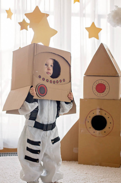 Sweet toddler boy, dressed as an astronaut, playing at home with cardboard rocket and handmade helmet from box - Photo, Image