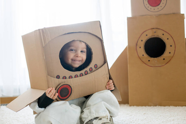 Sweet toddler boy, dressed as an astronaut, playing at home with cardboard rocket and handmade helmet from box - Foto, immagini