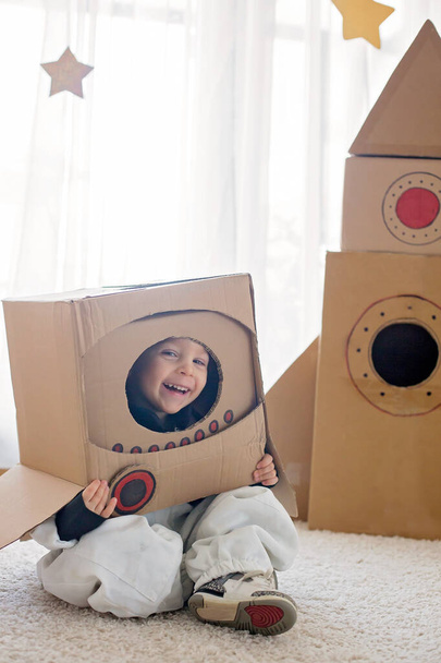 Sweet toddler boy, dressed as an astronaut, playing at home with cardboard rocket and handmade helmet from box - Foto, Imagem