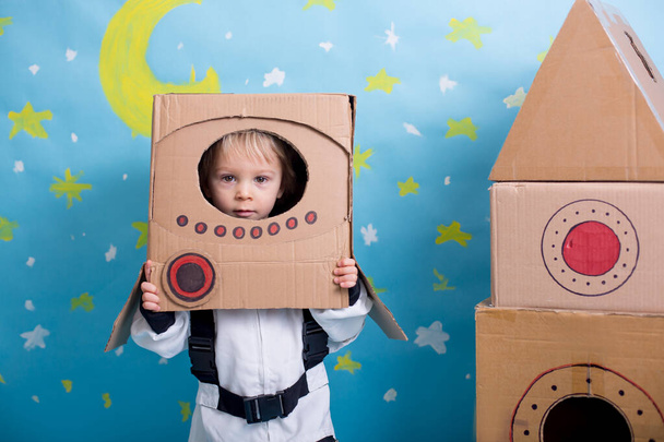 Sweet toddler boy, dressed as an astronaut, playing at home with cardboard rocket and handmade helmet from box - Foto, imagen