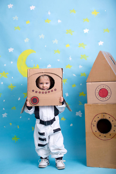 Sweet toddler boy, dressed as an astronaut, playing at home with cardboard rocket and handmade helmet from box - Фото, изображение