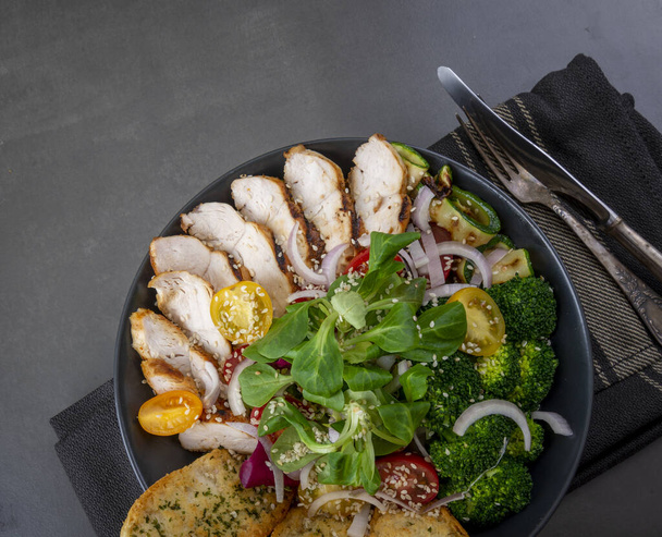 Salad of grilled chicken breast with broccoli, tomatoes and lamb's lettuce on a dark table. Variety of Caesar salad. Flat lay and copy space - Photo, Image