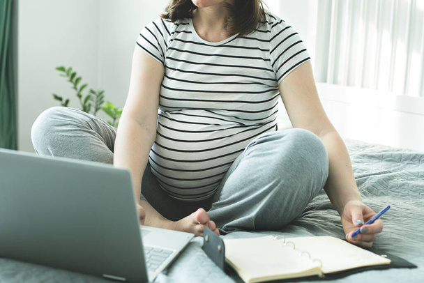 Young pregnant woman working from home office. Freelancer using laptop, notebook and the Internet. Workplace in bedroom. Concept of female business, career, home education, shopping online. - Photo, Image