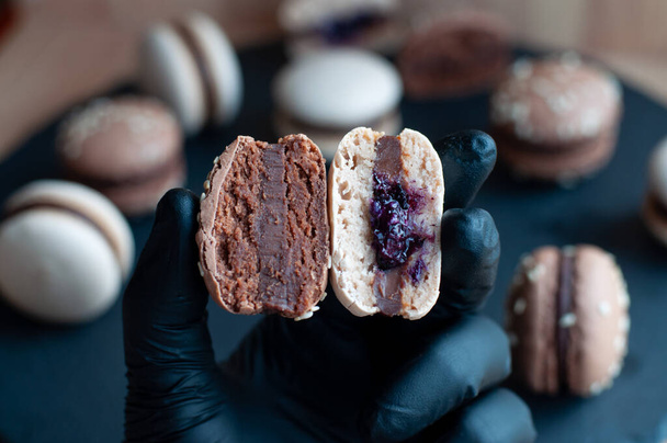 Mano en guantes negros sostiene dos mitades de macaron con ganache de chocolate
 - Foto, Imagen