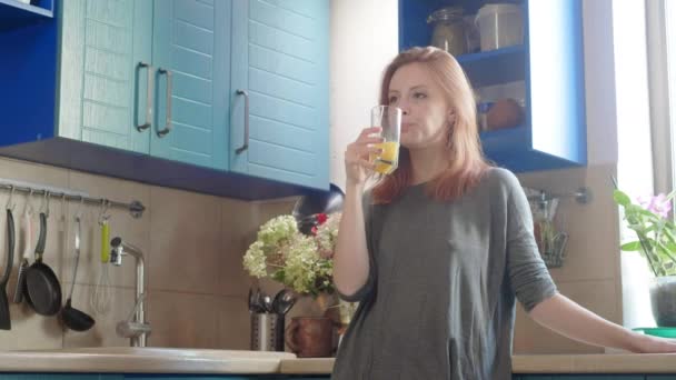 Beautiful girl drinking fresh orange juice from glass. Happy woman drinking fruit beverage. Pretty girl in t-shirt standing in kitchen. Healthy and natural nutrition. Healthy breakfast on the weekend - Filmagem, Vídeo