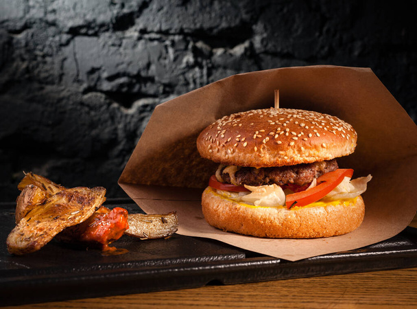 Burger und rustikale Kartoffeln auf einem Holztisch an einem Stand. Vor dem Hintergrund einer schwarzen Wand. - Foto, Bild