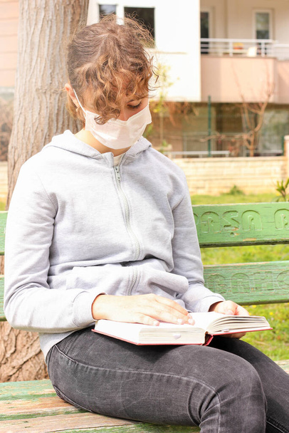 Young teenager girl with a medical mask reading a book in a park on bench. Quarantine and isolation during coronavirus - Foto, imagen