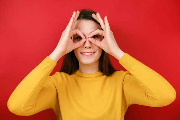 Cheerful funny young woman having fun making glasses shape with hands, does funny face with eyes wide open, dressed in yellow sweater, isolated on red studio background. People emotions concept - Foto, afbeelding