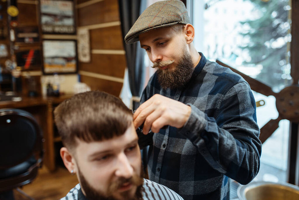 Barber drží hřeben a stříhá klientovi vlasy. Profesionální holičství je módní zaměstnání. Muž kadeřník a zákazník v retro stylu salonu - Fotografie, Obrázek