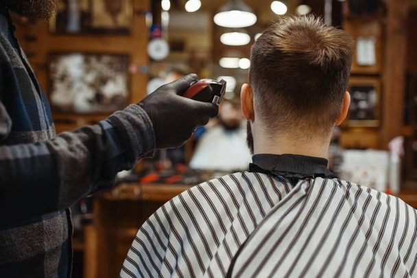 Barber in hat cuts the client 's hair. Professional barbershop is a trendy occupation. Male hairdresser and customer in retro style hair salon - Foto, Bild