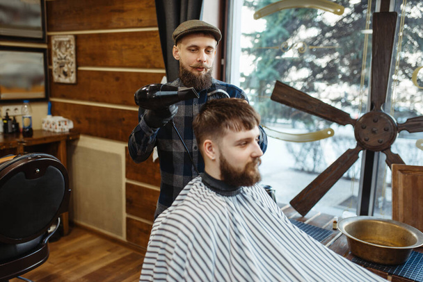 Barber with comb and hairdryer makes a hairstyle to a client. Professional barbershop is a trendy occupation. Male hairdresser and customer in retro style hair salon - Photo, Image