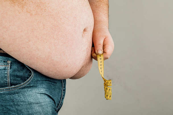 Un hombre mide su vientre gordo con una cinta métrica. sobre un fondo gris
 - Foto, imagen