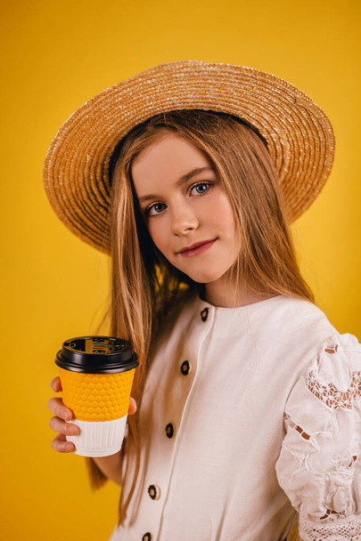 Young teenager girl holding a paper cup of coffee in her hands. On an isolated yellow background in a brown hat - Foto, Bild