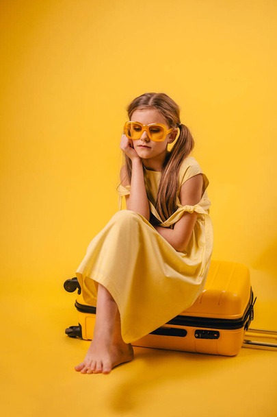Young tourist girl sitting on a suitcase in yellow glasses. Isolated on a yellow background. Travel concept. Flight Departure Canceled - Fotoğraf, Görsel