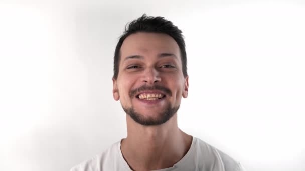 Blanke brunette man met kort haar met een baard in de studio op een witte achtergrond in een wit T-shirt glimlacht en lacht en gooit zijn hoofd naar achteren, close-up - Video