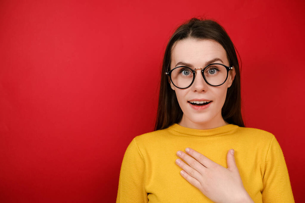 Joyous good looking young woman in glasses points hand at herself, can not believe her luck, has happy expression, wears yellow sweater, isolated over red studio background. Do you mean exactly me? - Photo, Image