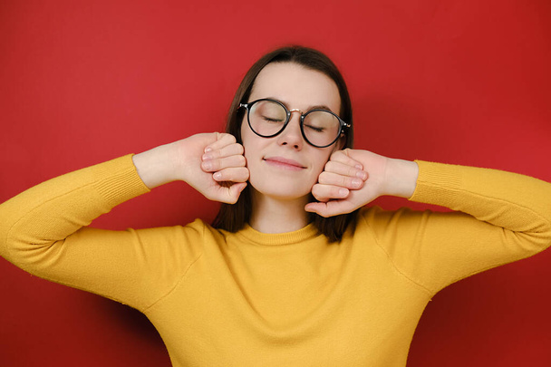 Happy woman in glasses smiles broadly, has sensual look, keeps hand near cheek, eyes closed, romantic expression, remembers something touching or pleasant, wears yellow sweater, isolated on red wall - Фото, зображення
