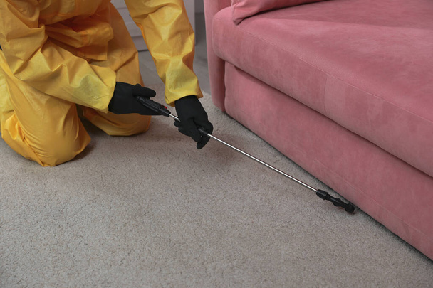 Trabajador de control de plagas en traje protector rociando insecticida bajo el sofá en interiores, primer plano
 - Foto, Imagen