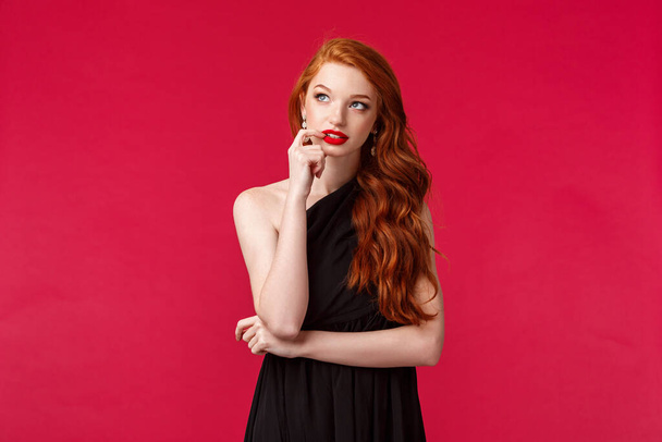 Portrait of thoughtful creative young redhead elegant woman with ginger hair in black dress, biting lip and looking up dreamy, thinking about solution, standing over red background - Photo, Image