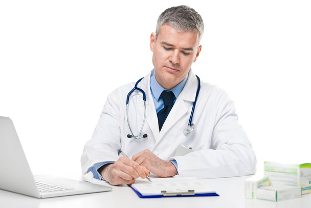 Lääkäri valkoisessa labratakissa istuu kirjoittamassa muistiinpanoja tai reseptiä toimistossaan keskittyen työhönsä valkoisella taustalla.
 - Valokuva, kuva
