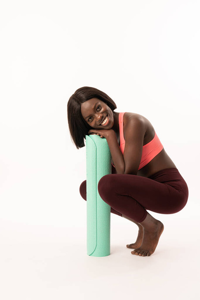 Jovem mulher africana feliz com tapete de ioga está pronto para a aptidão, sorrindo e olhando para a câmera, isolado sobre fundo branco
 - Foto, Imagem