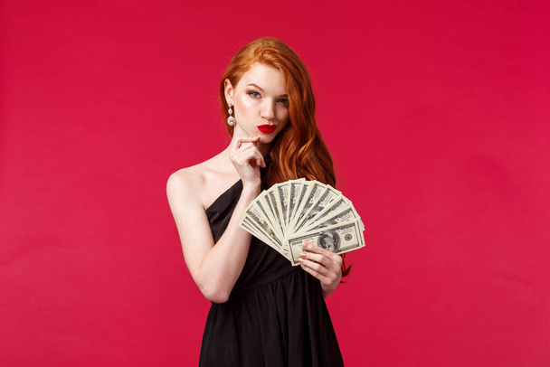 Luxury, beauty and money concept. Portrait of dubious and scheming good-looking young redhead woman in stylish black dress have interesting idea what buy on this cash dollars - Photo, Image