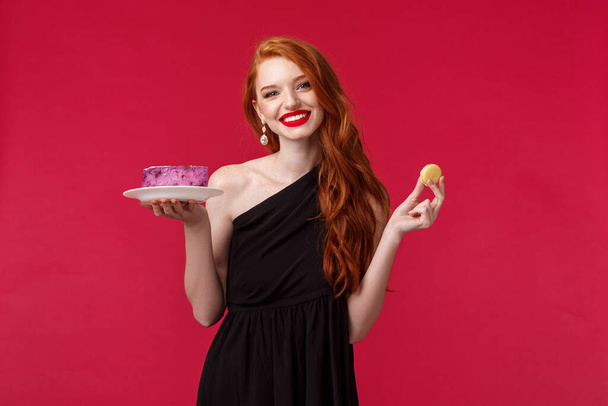 Elegant young woman in stylish black dress, red lipstick wearing black trendy dress over red background, holding biscuit and cake, smiling camera, cant resist temptation of desserts - Foto, Imagem