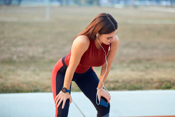 Nowoczesna młoda kobieta z telefonem komórkowym robi przerwę podczas joggingu / ćwiczeń. - Zdjęcie, obraz