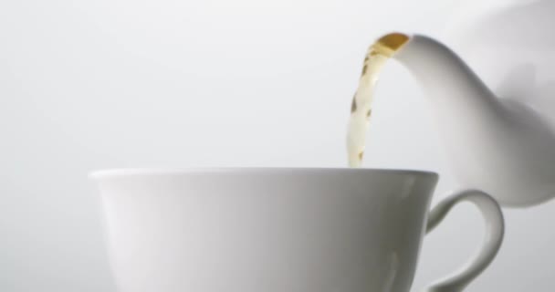Tea is poured from a white ceramic teapot into a white ceramic cup on a light background - Felvétel, videó