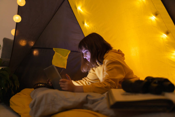 Pieni 11-vuotias tyttö käyttää kannettavaa tietokonetta kotitekoinen teltta sisällä olohuoneessa
. - Valokuva, kuva
