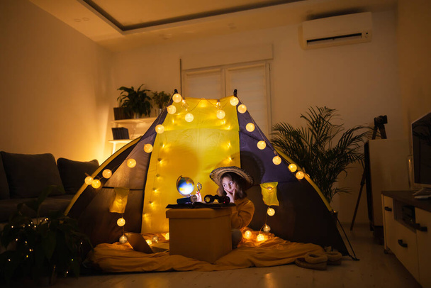Молодая девочка использует увеличительное стекло, чтобы исследовать земной шар в домашней гостиной палатке с легкими шарами
. - Фото, изображение