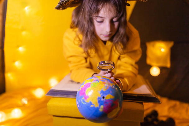 Young female child using magnifying glass to explore earth globe in a home made livingroom tent with light balls. - Photo, Image