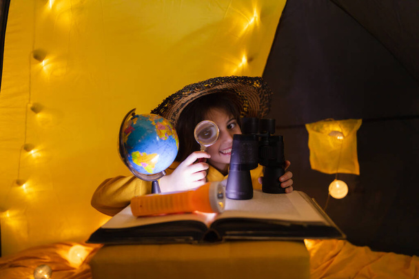 家の中で地球を探索するために虫眼鏡を使用して若い女性の子供作られたリビングルームのテントと光のボール. - 写真・画像
