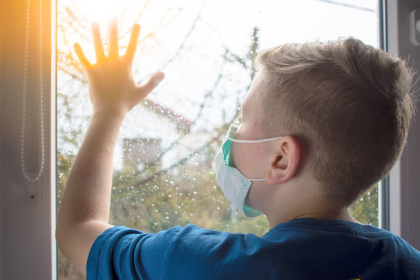 Een kind met een antiviraal masker. Het kind kijkt uit het raam.. - Foto, afbeelding