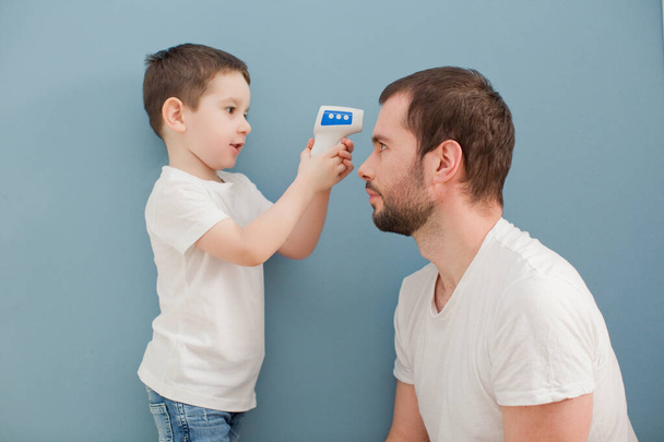 toddler boy is measuring his father's body temperature using infrared thermometer on a blue background - Foto, Imagem