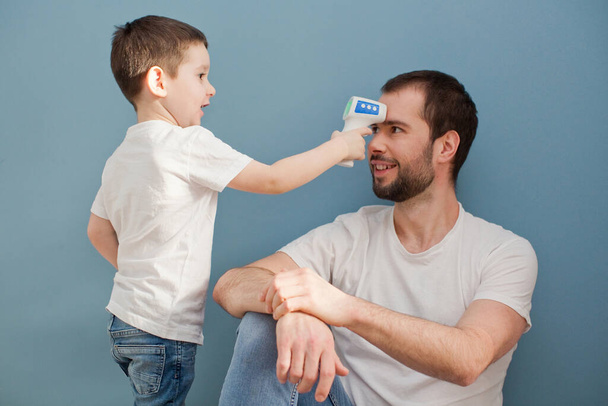 toddler boy is measuring his father's body temperature using infrared thermometer on a blue background - Fotó, kép