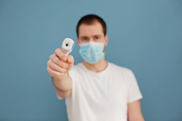joven con máscara quirúrgica está utilizando termómetro infrarrojo sobre un fondo azul
 - Foto, imagen