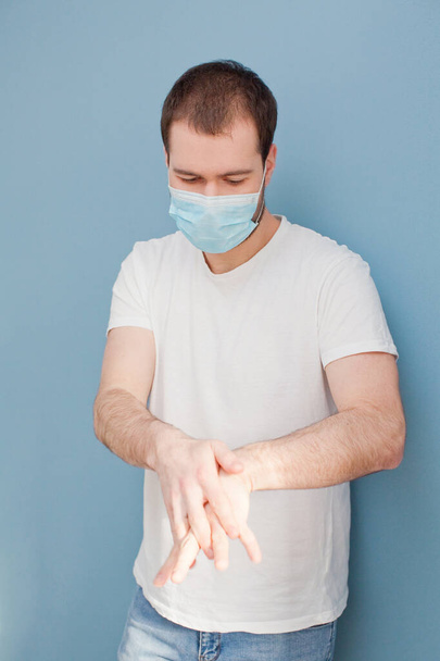 junger Mann mit OP-Maske reinigt seine Hände mit Händedesinfektionsmittel von Keimen auf blauem Hintergrund - Foto, Bild