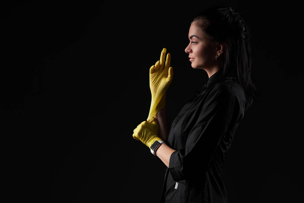 Young woman worker in black uniform is wearing protective yellow gloves. Dark studio background. Copy sapce for text - Zdjęcie, obraz