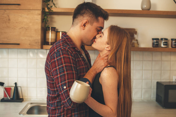 Schöne Ingwerfrau mit Sommersprossen küsst ihren Liebhaber, während sie in der Küche eine Tasse Tee trinkt - Foto, Bild