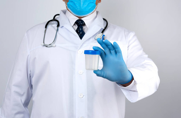 mężczyzna lekarz w białym płaszczu i krawacie stoi i trzyma pusty plastikowy pojemnik na próbkę moczu, w niebieskich jałowych rękawicach medycznych - Zdjęcie, obraz
