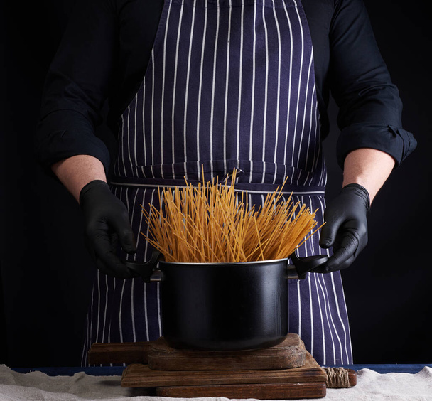 uncooked yellow long spaghetti in a black metal pan and a chef in a striped apron, cooking process - Fotografie, Obrázek