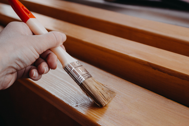 Γυναίκα ζωγράφος. Βουρτσίστε στο χέρι μιας γυναίκας, καλύπτοντας ξύλινες σανίδες και δοκούς με βερνίκι για εσωτερική εργασία στο χρώμα της βελανιδιάς. - Φωτογραφία, εικόνα
