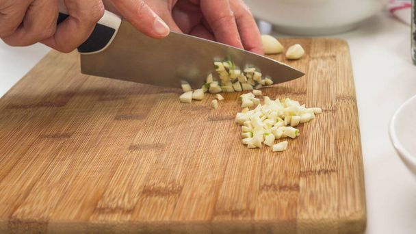 Picar el ajo en una tabla de cortar de madera, de cerca en una mesa de cocina, manos de mujer
 - Foto, Imagen