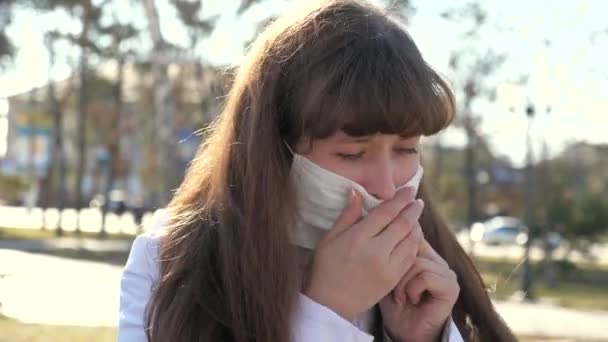 Egy fiatal beteg nő orvosi védőmaszkban köhög az utcán. A lány köhög. Fuvarozó, Covid-19, koronavírus, tünet. Pandémiás Coronavirus. egy influenzás ember. közelkép - Felvétel, videó