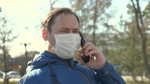 Προστασία από τον ιό του κερατοειδούς. Ο άνθρωπος με την ιατρική προστατευτική μάσκα κάθεται με ένα smartphone στο δρόμο της πόλης στην Ευρώπη. τουρίστες στο δρόμο φορούν προστατευτική μάσκα από ιούς. έννοια υγεία και ασφάλεια - Πλάνα, βίντεο