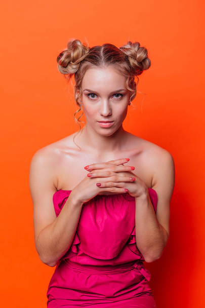 The girl in a red dress on a orange background in the studio. Blonde girl with two hair knots holding hands in front of her face and looking to the camera - 写真・画像